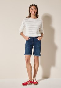 Suéter blanco de punto, pantalones cortos de mezclilla azules con dobladillo vuelto y zapatillas deportivas rojas con detalles blancos. El modelo está de pie con las manos en los bolsillos.