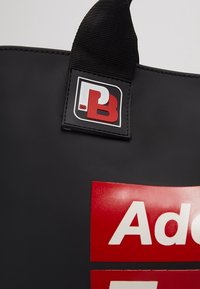 Sac fourre-tout noir avec une surface texturée, arborant un grand logo rouge et blanc sur le devant et une bandoulière noire avec une étiquette de marque.