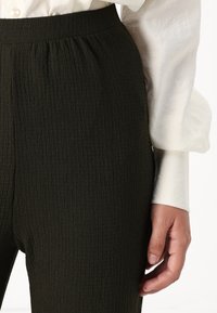 Dark green, textured pant with an elastic waistband. Close-up view shows subtle waves in the fabric and a hand resting at the side.