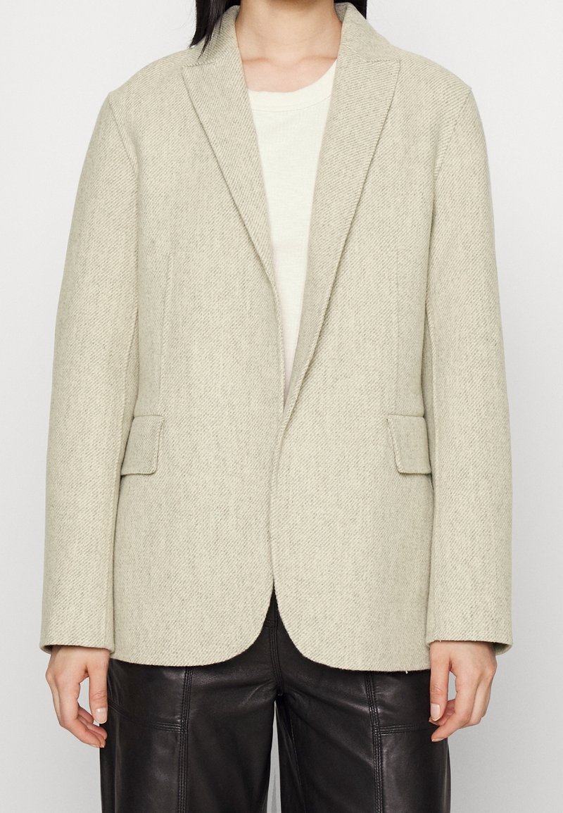 Svetle šedý blazer v herringbone vzoru vyrobený z texturované látky, s klopovými límci a dvěma předními kapsami, doplněný o krémový top.