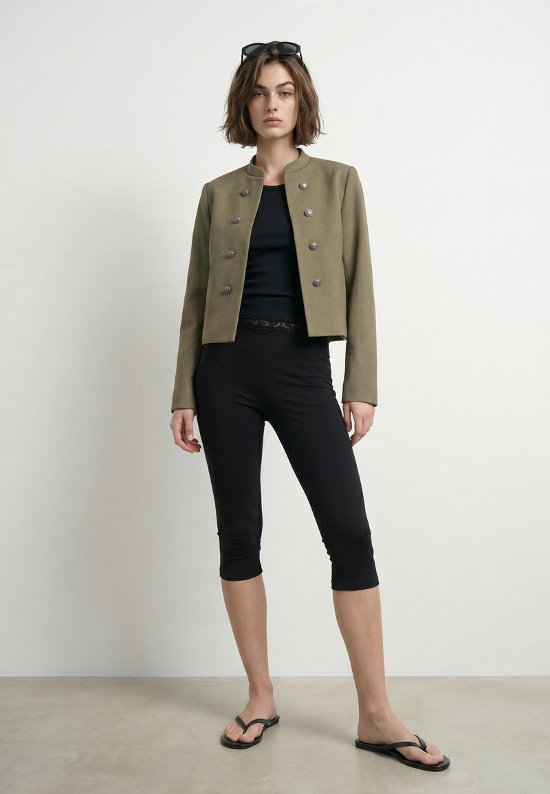 Femme debout portant une veste militaire vert olive, des leggings noirs courts, un top noir, des tongs noires et des lunettes de soleil sur la tête.