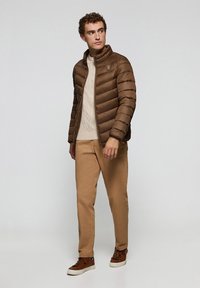 Chaqueta acolchada marrón con costuras horizontales, cuello alto y un parche de logo. Combinada con un suéter de punto en color crema y pantalones beige. Zapatillas marrones.