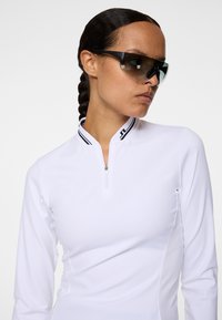 Haut blanc à manches longues avec un col demi-fermeture éclair présentant des accents rayés noirs et blancs ; texture lisse avec une silhouette ajustée. Lunettes de soleil noires incluses.