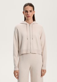 Felpa corta di colore beige chiaro con zip, realizzata in tessuto morbido, dotata di cappuccio foderato, polsini a coste e una texture liscia.