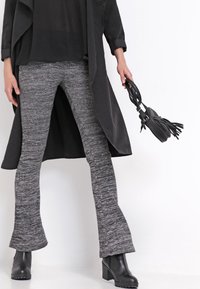 Manteau long noir sur un haut noir, associé à un pantalon évasé texturé gris. Bottines noires à talons et un petit sac noir avec des franges.