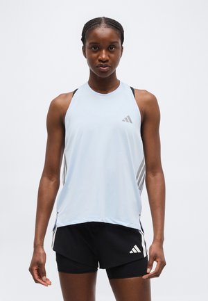 Αθλητική γυναίκα που φοράει αμάνικη ανοιχτό μπλε μπλούζα Adidas και μαύρο σορτς Adidas, μπροστά σε απλό λευκό φόντο.