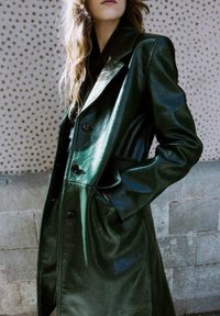 Cappotto lungo in pelle verde con finitura lucida, grandi rever e bottoni, dotato di tasche laterali e silhouette strutturata.