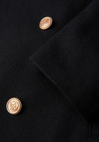 Manteau en laine noire avec une texture lisse, un col cranté et des boutons dorés au design complexe. Vue rapprochée.