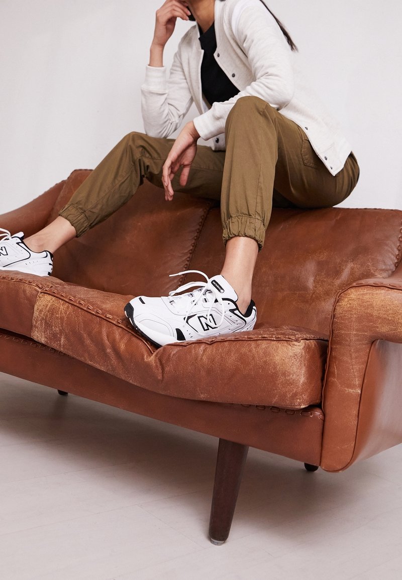 Personne portant des baskets blanches, un pantalon de jogging marron et une veste blanche assise sur un canapé en cuir marron usé.
