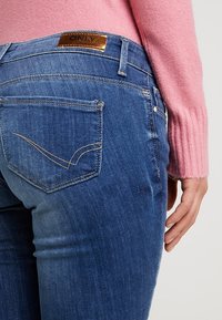 Närbild av en person som bär blå denimjeans med en brun "ONLY"-etikett och en rosa långärmad tröja, handen vilande vid sidan.