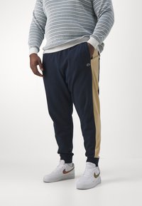 Calças de jogging em azul-marinho com painéis laterais em tom claro, cintura elástica e bolsos. Usadas com ténis brancos com um swoosh em bronze.