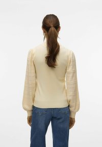 Vrouw met lang bruin haar in een lage paardenstaart draagt een crèmekleurige gebreide trui met textuur en pofmouwen, en spijkerbroek, staande tegen een effen achtergrond.