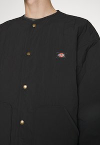 Veste matelassée noire avec boutons-pression, poches à l'avant et une petite écusson avec le logo Dickies sur la poitrine gauche, portée par une personne.