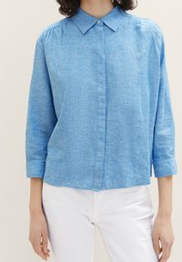Chemise bleu clair à col en tissu texturé, avec un devant boutonné et des manches trois-quarts, associée à un pantalon blanc.