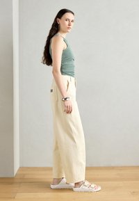Pantaloni larghi beige chiaro, top in tessuto verde menta, sandali bianchi con fibbie dorate e un braccialetto d'argento. Modello in posa di lato.