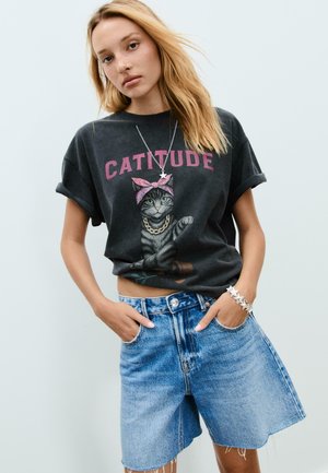 Black graphic t-shirt featuring a cat with a pink bandana and "CATTITUDE" text, paired with light blue denim shorts and silver star bracelet.