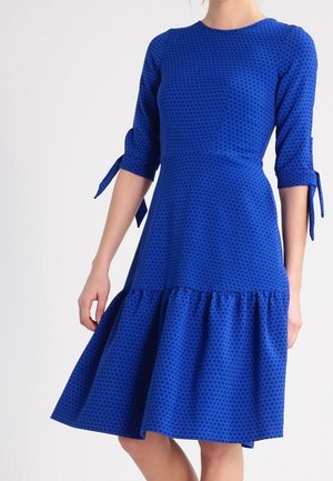 Frau trägt ein knielanges blaues Kleid mit schwarzen Punkten, dreiviertel langen Ärmeln mit Schleifen und einem ausgestellten Rock.