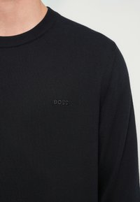 Μαύρο πλεκτό πουλόβερ με στρογγυλή λαιμόκοψη, με διακριτικό τόνο "BOSS" στο στήθος. Λείο υφή και εφαρμοστή σχεδίαση.
