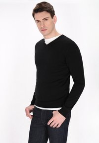 Pullover nero a V in maglia realizzato in un tessuto morbido, con maniche lunghe e design aderente, indossato sopra una T-shirt bianca e abbinato a jeans scuri.