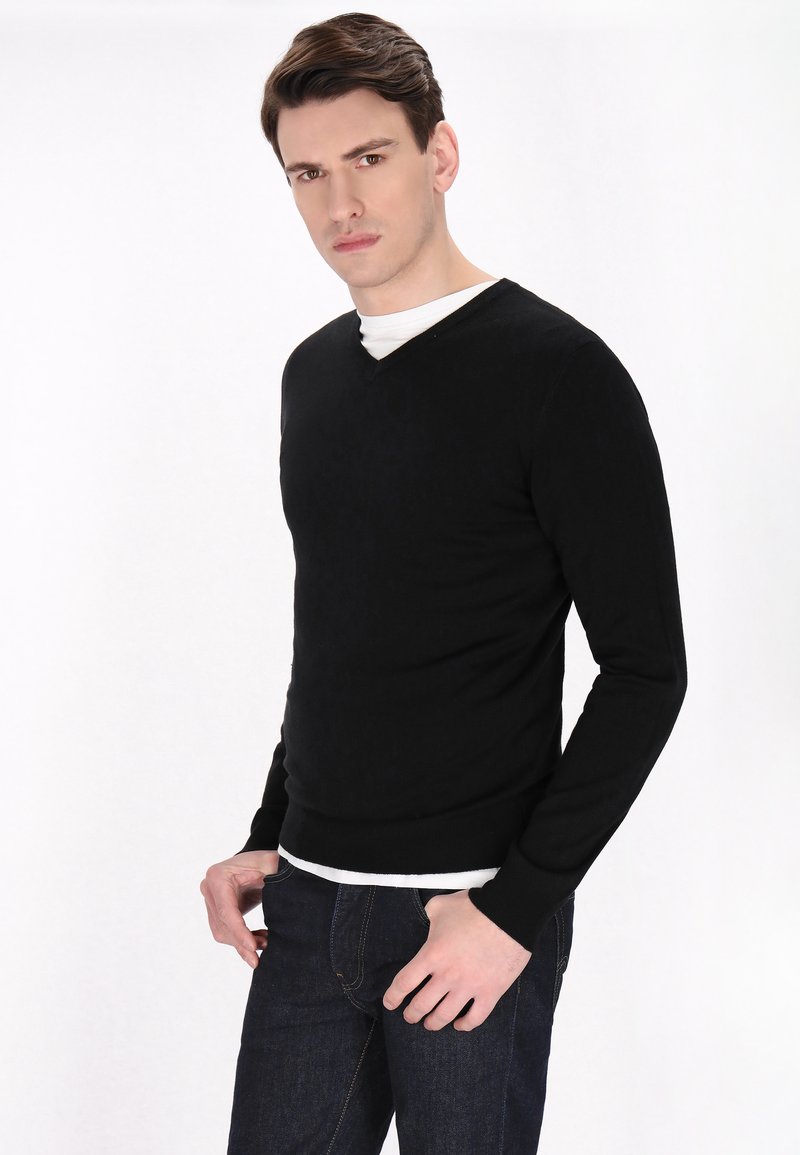 Pullover nero a V in maglia realizzato in un tessuto morbido, con maniche lunghe e design aderente, indossato sopra una T-shirt bianca e abbinato a jeans scuri.