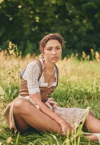 Vêtement traditionnel avec un corsage marron à manches en dentelle, une jupe rose avec broderie et un lien en ruban, dans un champ herbeux.