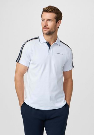 Man in een wit polo met korte mouwen en schouderstrepen en donkerblauwe broek, staand met handen in de zakken, kijkend naar rechts.