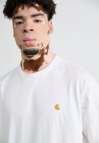 T-shirt en coton blanc avec un petit logo doré brodé sur la poitrine. Encolure ronde et texture lisse, porté par une personne.