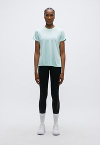 Αθλητική κοντομάνικη μπλούζα σε ανοιχτό τιρκουάζ χρώμα, συνδυασμένη με μαύρο κολάν που έχει διάφανα πάνελ, άσπρες αθλητικές κάλτσες και άσπρα παπούτσια.