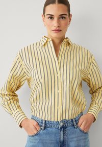 Chemise boutonnée à rayures jaunes avec des lignes verticales bleu marine, dotée d'un col et de manches longues, associée à un jean en denim bleu clair.