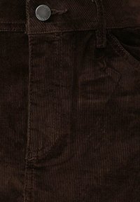 Bruine corduroy stof, met verticale ribbels, een zilveren knoopsluiting en zijzakken. Gestructureerd oppervlak met een subtiele glans.