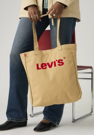 Osoba ubrana w niebieskie dżinsy i czarne buty trzyma beżową torbę na ramię Levi's z czerwonym logo, stojącą obok czerwonego i chromowanego krzesła.