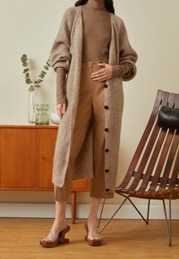 Kvinna som bär en lång beigefärgad kofta, brun polotröja, korta bruna byxor och klacksandaler med klara remmar, stående i ett rum med mid-century stil.