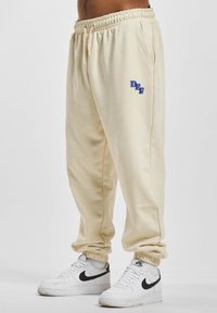 Beige sporta bikses ar elastīgu jostu, sānu kabatām un zilu "DFF" logo uz kreisās augšstilba. Izgatavotas no mašīnā adīta auduma, ar sašaurinātām piedurknēm.