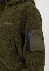 Oliwkowa polarowa kurtka z teksturowanym, miękkim wykończeniem. Posiada zamek błyskawiczny na kieszeni na ramieniu, z gładką tkaniną i subtelnym obszyciem.