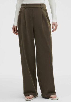 Pantaloni largi kaki cu pliuri, purtați cu un top alb cu mâneci lungi și sandale albe tip flip-flop.