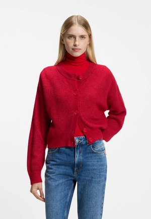 Kvinde med lyst, glat hår iført en rød højhalset trøje og rød cardigan med knapper, kombineret med blå jeans, står foran en hvid baggrund.