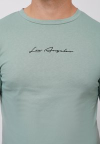 Camiseta de manga corta verde menta con cuello redondo y texto en cursiva negro "Los Ángeles" en el pecho. Textura de tela suave y lisa.