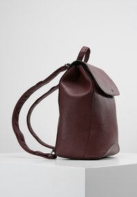 Zaino in pelle bordeaux con chiusura a patta, parte superiore piatta e spallacci regolabili. Presenta una texture liscia e un design del corpo arrotondato.