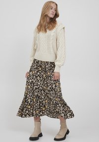 Pullover spesso a maglia a treccia, gonna plissettata nera a fiori con dettagli gialli e stivaletti beige con suola chunky.