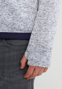 Närbild av en hand som delvis är täckt av en ljusgrå prickig stickad långärmad tröja med tumhål, vilande nära fickan på mörkgrå byxor.
