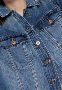 Denim jacket in mid-blue with chest pockets, button closure, pointed collar, and subtle stitching details. Soft texture with a classic fit.