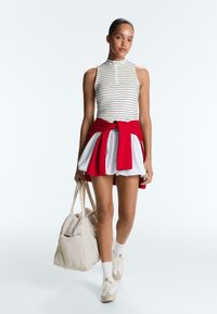 Femme portant un haut rayé sans manches et une jupe blanche avec un pull rouge noué autour de la taille, portant un sac beige, marchant vers l'avant sur fond blanc.