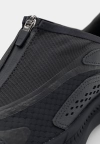 Chaussure noire à enfiler avec fermeture éclair, tissu texturé et semelle en caoutchouc avec des accents perforés pour la respirabilité.