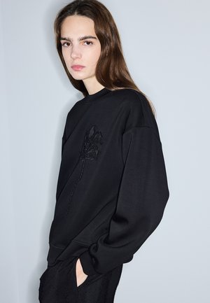 Jonge vrouw met lang bruin haar draagt een zwarte sweatshirt met een subtiel bloemenpatroon, staand tegen een lichtgrijze achtergrond, handen in de zakken.