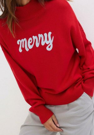 Femme portant un pull rouge avec un texte blanc « merry » et un pantalon gris clair, les mains détendues le long du corps sur un fond uni.