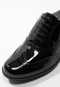 Chaussure à lacets en cuir verni noir avec un design brogue, une texture lisse, un bout arrondi et des perforations décoratives le long de la tige.