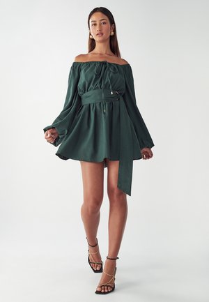 Zielona sukienka typu off-shoulder wykonana z miękkiego materiału, z wcięciem w talii i wiązaniem oraz zwiewnymi, długimi rękawami. Widoczne akcenty ze złotego metalu.