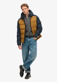 Pufjakke i marineblå og gyldenbrun, med lynlås foran, parret med en grå pullover, lyseblå jeans og mørke snørebåndsstøvler.