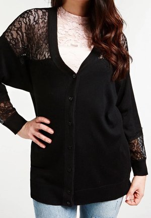 Femme portant un cardigan noir boutonné avec des manches et épaules en dentelle par-dessus un haut en dentelle rose clair et un jean bleu clair.