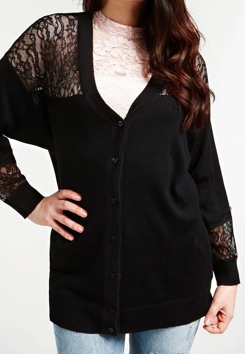 Femme portant un cardigan noir boutonné avec des manches et épaules en dentelle par-dessus un haut en dentelle rose clair et un jean bleu clair.